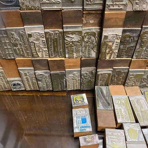 May include: A collection of vintage metal printing blocks with wooden bases. The blocks feature various designs, including text, illustrations, and logos. Some blocks are stacked, while others are arranged on a wooden surface.