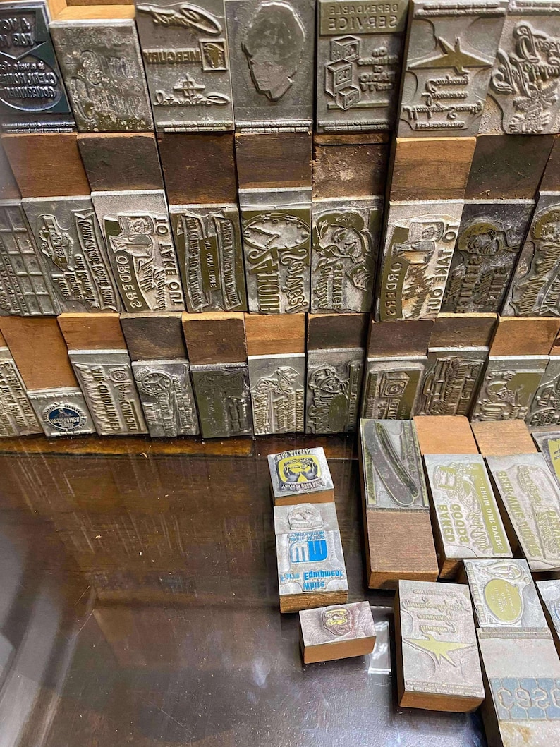 May include: A collection of vintage metal printing blocks with wooden bases. The blocks feature various designs, including text, illustrations, and logos. Some blocks are stacked, while others are arranged on a wooden surface.
