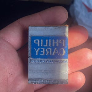May include: A rectangular metal printing block with the text "PHILIP CABREA" in white, set against a blue background. Additional text is visible below. The block is held in a hand.