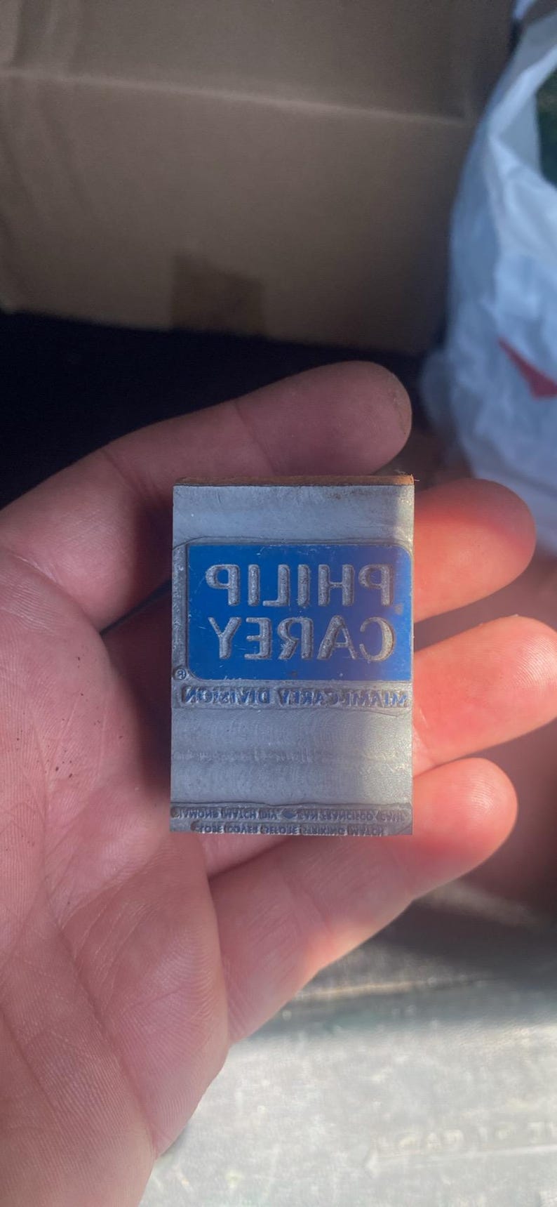May include: A rectangular metal printing block with the text "PHILIP CABREA" in white, set against a blue background. Additional text is visible below. The block is held in a hand.