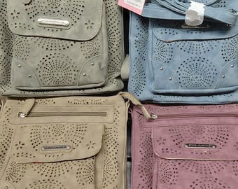 May include: Four crossbody bags in gray, blue, tan, and mauve. Each bag features a front flap with laser-cut circular patterns, small silver studs, a zippered top compartment, and a rectangular metal logo plate labeled Stone Mountain USA.