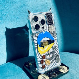 Puede incluir: Funda transparente para teléfono con un diseño único que presenta labios azules y amarillos, una bala, granos de café y detalles decorativos. La funda tiene adornos negros en forma de cuerno en la parte superior. La funda está sobre un soporte negro.