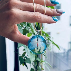 May include: A round pendant necklace with a blue and white flower pressed inside. The pendant is on a cream-colored cord.