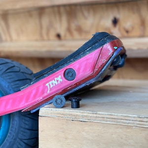 Könnte beinhalten: Ein rotes und schwarzes elektrisches Skateboard mit dem Wort "Jinx" in weißen Buchstaben. Zwei schwarze zylindrische Komponenten befinden sich vor dem Skateboard. Das Skateboard hat einen schwarzen Reifen und ein blaues Rad.