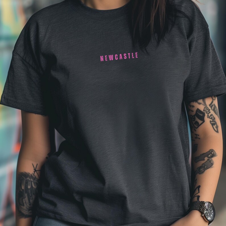 K&ouml;nnte beinhalten: Ein dunkelgraues T-Shirt mit dem Wort "NEWCASTLE" in Pink auf der Vorderseite.