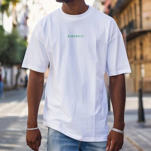 K&ouml;nnte beinhalten: Wei&szlig;es Oversize-T-Shirt mit gr&uuml;nem "NEWCASTLE"-Schriftzug auf der Vorderseite. Das Model tr&auml;gt eine blaue Jeans.