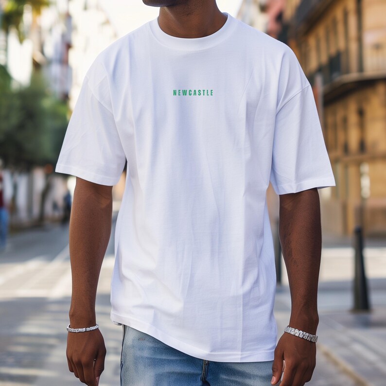 K&ouml;nnte beinhalten: Wei&szlig;es Oversize-T-Shirt mit gr&uuml;nem "NEWCASTLE"-Schriftzug auf der Vorderseite. Das Model tr&auml;gt eine blaue Jeans.