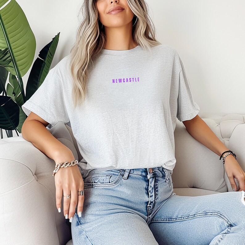 K&ouml;nnte beinhalten: Ein graues T-Shirt mit dem Wort "NEWCASTLE" in Lila auf der Vorderseite. Das Shirt wird von einer Person mit langen blonden Haaren und einer blauen Jeans getragen.