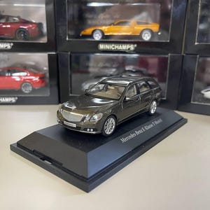 Puede incluir: Un modelo a escala de un Mercedes-Benz E-Class T-Modell en color gris oscuro, expuesto sobre una base negra. El coche de juguete tiene ruedas plateadas. Otros modelos de coches se ven en el fondo.