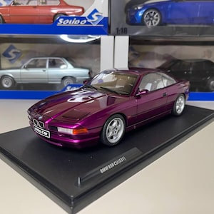 Puede incluir: Un modelo de coche BMW 850 CSi (E31) de color púrpura metálico sobre un soporte de exhibición negro. El modelo tiene ruedas plateadas y un interior detallado. El coche se exhibe frente a otros modelos de coches en su embalaje original.