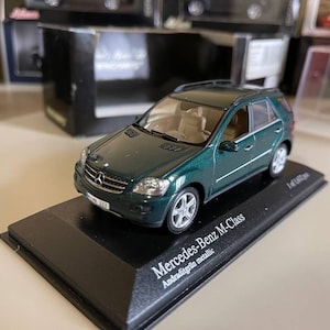May include: A dark green Mercedes-Benz M-Class model car on a black display base. The car has a black interior and silver wheels. The base has the text "Mercedes-Benz M-Class Andaragitgrün metallic" printed on it.