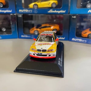 May include: A 1/18 scale diecast model car of a BMW 320i race car, finished in yellow and white. The car features a black and red KW suspension sticker on the front bumper. The car is displayed on a black base with the text "BMW 320i" printed on it.