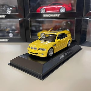 May include: A yellow 1/18 scale diecast model car of a BMW M3 Coupe. The car is on a black display base with the words "BMW M3 Coupe" printed on it.