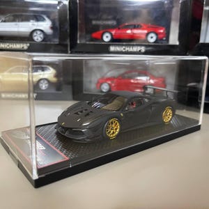 Pode incluir: Um carro modelo preto fundido com rodas douradas é exibido em uma caixa de acrílico transparente. O modelo é um carro esportivo com detalhes. Outros modelos de carros são visíveis ao fundo.