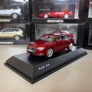 以下が含まれることがあります： 黒い展示台に置かれた赤いアウディA4の模型。 模型車は、銀色のホイールとナンバープレートが特徴の精巧なレプリカです。 展示台には「Audi A4」と表示されています。 他の模型車も背景に見えます。