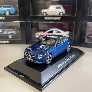 Könnte beinhalten: Ein blaues Mercedes-Benz C-Klasse Modellauto auf einem schwarzen Präsentationsständer. Das Miniaturauto hat eine glänzende Oberfläche und detaillierte Merkmale, darunter silberne Räder und ein schwarzes Dach. Der Präsentationsständer trägt den Namen des Autos.