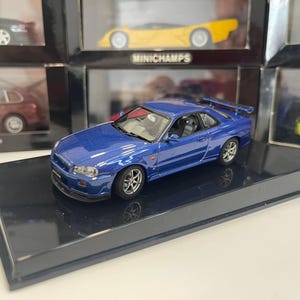 Puede incluir: Un coche de juguete de metal azul estilo cupé con alerón trasero y ruedas plateadas, expuesto sobre una base rectangular negra. El vehículo tiene ventanas transparentes, faros detallados y un interior visible con asientos de tipo baquet.