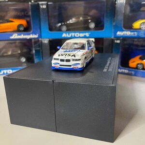 May include: A white and blue 1/18 scale model car with the words "Round Wisa" on the side. The car is on a black display stand.