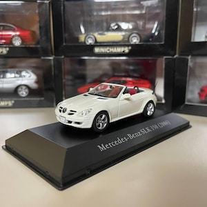 Puede incluir: Un modelo de coche blanco Mercedes-Benz SLK 350 (2006) con interior rojo, expuesto sobre una base negra. El modelo forma parte de una colección de coches en miniatura, con otros vehículos visibles al fondo.