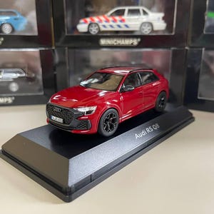 Op de afbeelding: Een rood Audi RS Q8 modelauto op een zwarte displaystandaard. De modelauto heeft zwarte wielen en een gedetailleerde grille. De displaystandaard heeft de tekst "Audi RS Q8" erop gedrukt. Andere modelauto's zijn zichtbaar op de achtergrond.