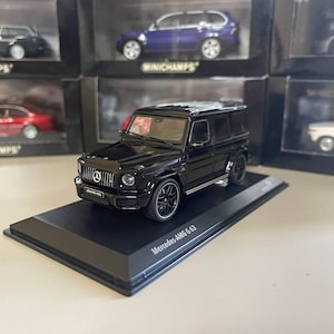 Puede incluir: Modelo de coche de fundición a presión Mercedes-AMG G 63 negro sobre una base de exhibición negra. El coche tiene ruedas negras y un interior negro. La base tiene el texto "Mercedes-AMG G 63" impreso en ella.