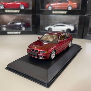 May include: A burgundy BMW model car, approximately 6 inches long, sits on a black display stand. The miniature car has a detailed interior and silver wheels. Other model cars are visible in the background.