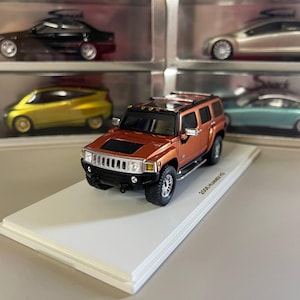 Può includere: Un modello in scala 1/18 di un Hummer H2 arancione del 2003. Il modello è su una base di esposizione bianca con il testo "2003 HUMMER H2" stampato su di essa.