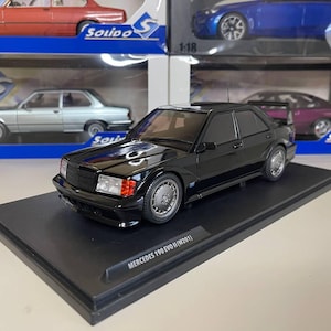 Puede incluir: Un coche modelo negro Mercedes 190 EVO II (W201) sobre un expositor negro. El coche modelo es una réplica detallada con ruedas plateadas y luces traseras rojas. El expositor tiene el texto "MERCEDES 190 EVO II (W201)". Otros modelos de coches son visibles en el fondo.