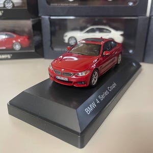 May include: A red BMW 4 Series Coupe model car on a black display stand. The model car has detailed features, including a sunroof and silver wheels. The display stand has the text "BMW 4 Series Coupe" in white.