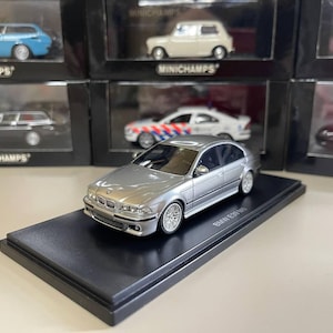 May include: Silver BMW E39 M5 diecast model car on a black display stand. The model is highly detailed with realistic features. Other model cars are visible in the background, displayed in their original packaging.