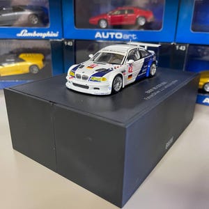 May include: A white, blue, and black BMW M3 GTR model car with the number 42 on the side, displayed on a black box. Other model cars are visible in the background.