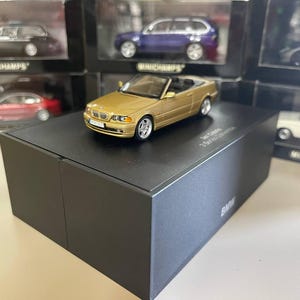 May include: A gold 1:18 scale model of a BMW 3 Series convertible. The model is on a black display box with the BMW logo.