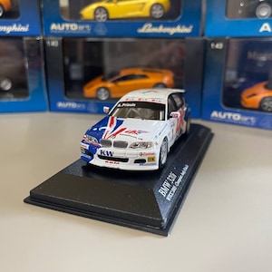 May include: A white and blue BMW 320i race car model with the text "WTCC 2005 Champion" on the base. The car has a red, white, and blue livery with the number "3" on the side.