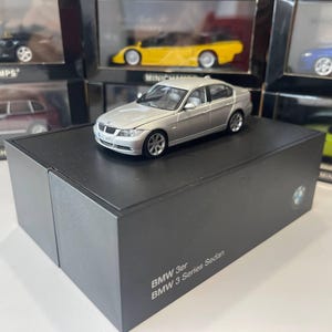 May include: A silver BMW 3 Series sedan die-cast model car displayed on a black rectangular box. The box features the text BMW 3er and BMW 3 Series Sedan printed in white. The model car has detailed headlights, a front grille, and wheels.