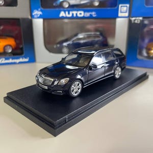 May include: A dark blue diecast model car of a station wagon, displayed on a black rectangular base. The model has silver wheels and detailed features. Other model cars are visible in the background, in their original packaging.