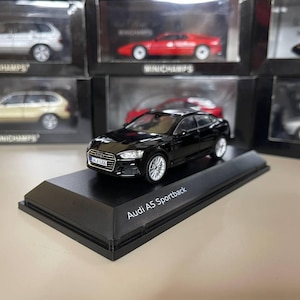Könnte beinhalten: Ein schwarzes Audi A5 Sportback Modellauto auf einem schwarzen Präsentationsständer. Das Modell hat silberne Räder und detaillierte Scheinwerfer. Weitere Modellautos sind im Hintergrund zu sehen.