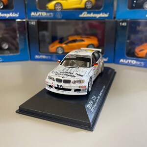 May include: A white 1/43 scale model car of a BMW 320i race car with the text "Original BMW Teile und Zubehör" and "KW" on the side. The car is on a black display base with the text "BMW 320i ETCC 2005" printed on it.