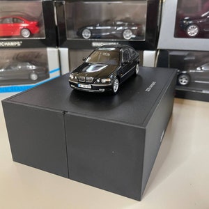 May include: A black diecast model car of a BMW 325ti compact, displayed on a black box. The model has silver wheels and detailed headlights. Other model cars are visible in the background, some in their original packaging.