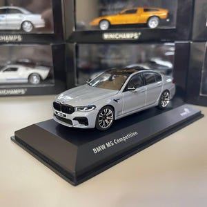 Peut inclure: Une voiture miniature BMW M5 Competition argentée sur un socle noir. La voiture miniature a un toit noir et des roues détaillées. Le socle porte l'inscription "BMW M5 Competition". D'autres voitures miniatures sont visibles en arrière-plan.