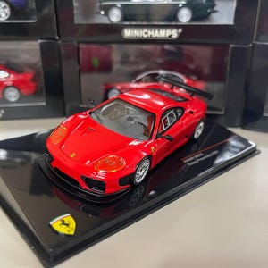 Op de afbeelding: Een rode Ferrari 360 Modena diecast model auto op een zwarte display basis. De auto heeft een zwarte achtervleugel en zwarte wielen. De basis heeft het Ferrari logo en de tekst "Minichamps" erop gedrukt.