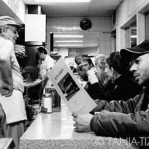 May include: Black and white photograph of people in a restaurant. A man wearing a black cap is seated at a table with a menu. He is looking at the camera. There are other people in the background.