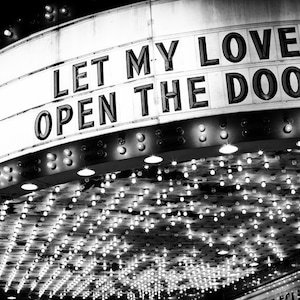 May include: Black and white photo of a vintage marquee sign with the text "LET MY LOVE OPEN THE DOOR" in white letters. The sign is lit with many small light bulbs.