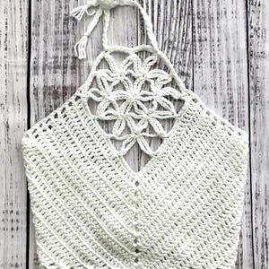 May include: A white crochet halter top with a floral design on the chest. The top features a V-neck and a scalloped edge. The halter straps are tied at the back of the neck.
