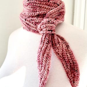 May include: A hand-knitted scarf in shades of pink, with flecks of black and white. The scarf is tied in a knot at the neck, with the ends hanging down. The scarf is made of a soft, textured yarn.