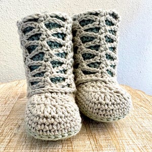 May include: A pair of handmade crochet boots in a neutral beige color with a lattice design on the upper shaft. The interior is a darker green. The boots are sitting on a wooden surface.