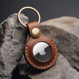May include: A brown leather AirTag holder with a gold-coloured key ring and button clasp. The holder has a stitched border and a circular window displaying the Apple logo. The background is a dark grey.