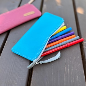 Op de afbeelding: Een felblauw etui met rits en een witte rits, gevuld met kleurrijke stiften. Het etui is gemaakt van een getextureerd lederachtig materiaal.