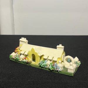 May include: A miniature yellow cottage figurine with a cream-colored roof and chimneys. The cottage has a small arched doorway and is surrounded by colorful miniature bushes and flowers. A small white fence and a circular window are also present.