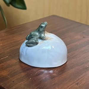 May include: A small ceramic figurine of a green frog sitting on a white, rounded base. The frog is looking up and has a detailed texture.