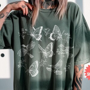May include: An olive green t-shirt featuring a white illustration of butterflies, birds, and floral elements. The shirt has a relaxed fit and a vintage aesthetic. The text "MOSS" is visible on the bottom right.